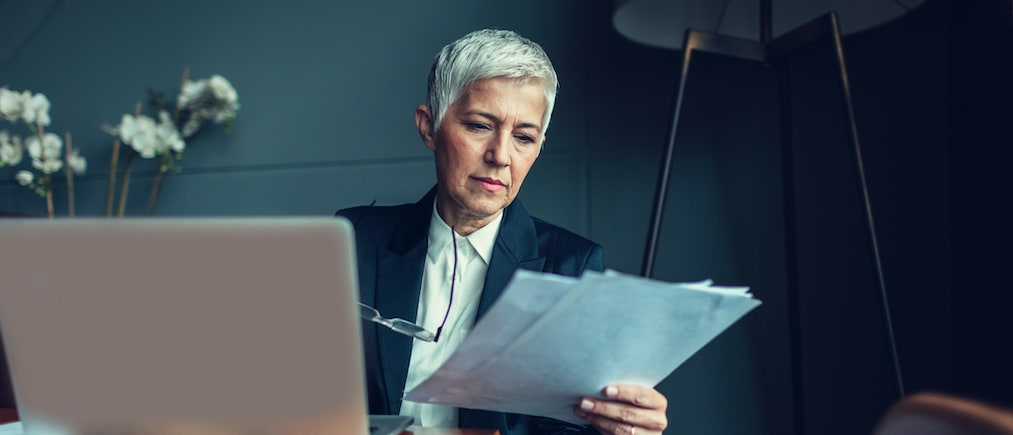Portrait of mature businesswoman, sitting in her office and finishing paperwork. Holding eyeglasses. In front of her on the desk is laptop.