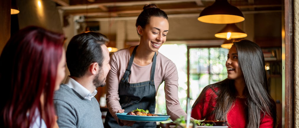 Happy waitress serving food to a group of friends having dinner  at a restaurant - food service concepts 