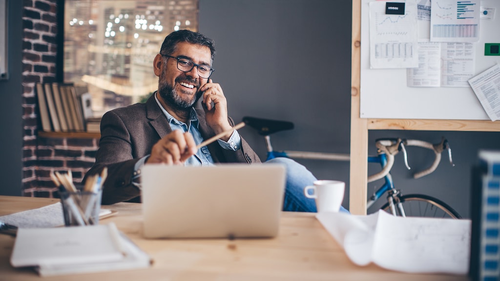 Mature business man working at modern office space, working on project. Making a call on mobile phone at his desk.  Brick wall and bicycle in  background.